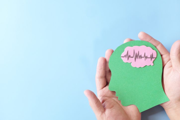 Two hands hold a green paper cutout of a human head with a pink brain and ECG line inside, set against a light blue background.