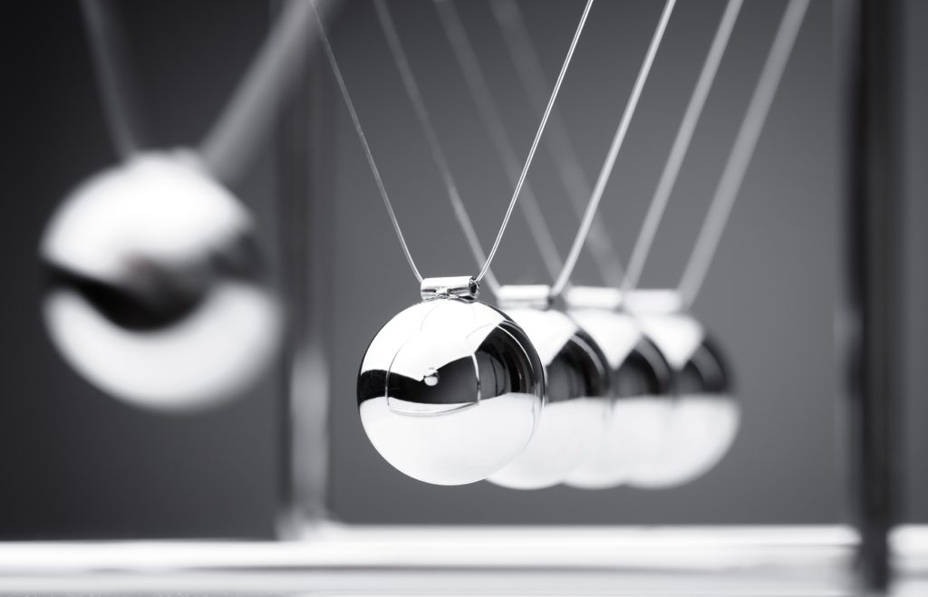 Photo of a Newton’s cradle with five shiny metallic spheres suspended by thin wires, showing one sphere pulled back and the others aligned in a row. The background is gray and out of focus, emphasizing the polished reflective surfaces of the spheres.
