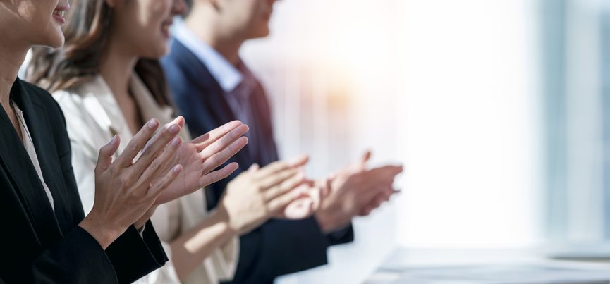 Three people dressed in business attire are standing in a line while smiling and clapping. 