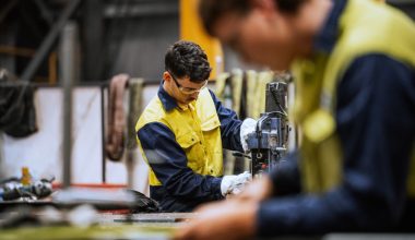 Two individuals in yellow and blue work uniforms in an industrial workshop; one operates a machine while the other is blurred in the foreground, surrounded by tools and equipment.