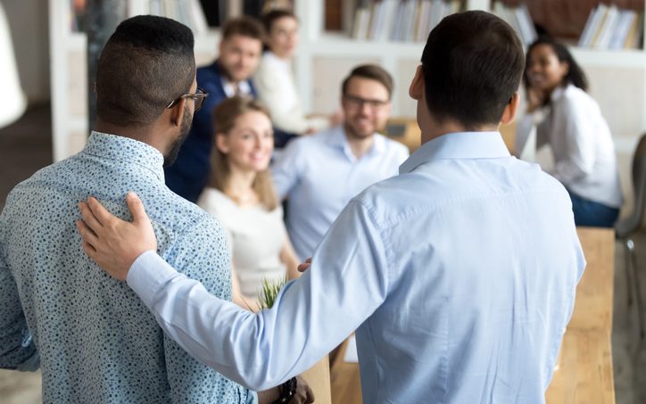 Two men are standing with their backs to the camera as they face a group of six men and women who are sitting. One of the men who is standing is introducing the other to the group. 