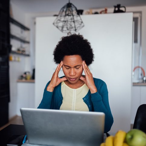 A woman is sitting at a table in a kitchen with an open laptop in front of her and her head in her hands. 