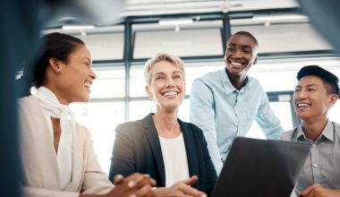 A diverse group of four professionals in business attire, smiling and laughing together while gathered around a laptop.