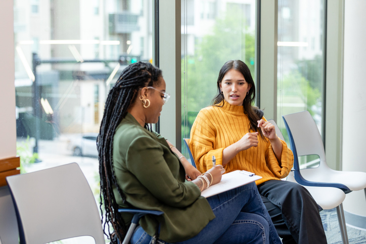 Two women sitting in chairs having a conversation while one takes notes on a clipboard. 