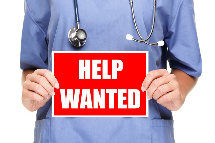 Person in medical scrubs holding a "HELP WANTED" sign.