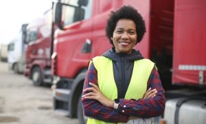 A black woman is wearing a safety vest and standing in front of a row of large parked trucks.
