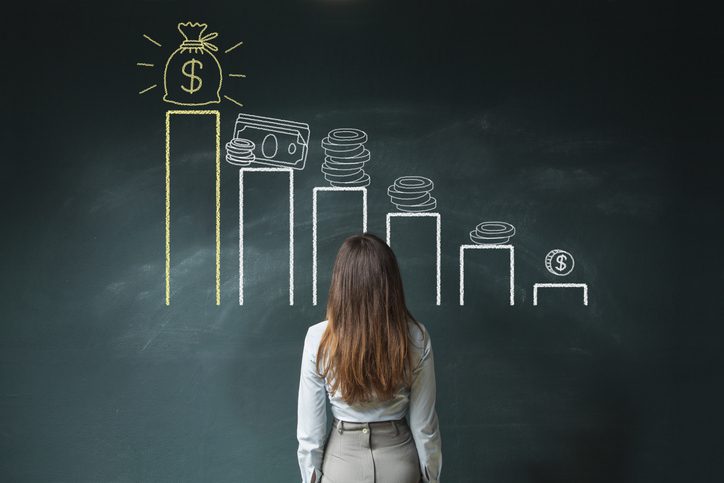 A woman stands in front of a blackboard with an illustrated bar graph showing an upward trend, each bar topped with various forms of money.