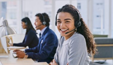 Smiling woman in a headset at an office desk, with two colleagues in the background.