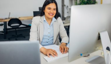 Woman typing on a keyboard in an office setting, with a large monitor in front of her.