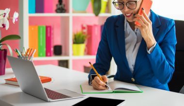 Person in a blue blazer talking on a phone and writing at a desk with a laptop and tablet.