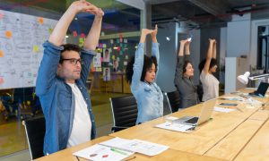 Four people sitting at a conference table in an office, stretching their arms upwards.
