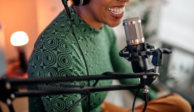 A Black woman in a green patterned shirt wearing headphones and speaking into a microphone in a recording setting.