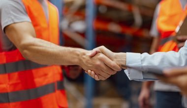 Photo of two people shaking hands in a warehouse. One man wears an orange safety vest and gray shirt; the other person wears a light blue long‑sleeve shirt. Shelving and another person in an orange vest are visible in the background.