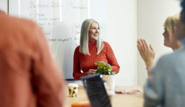 Photo of a woman with long gray hair wearing a red turtleneck sweater standing in front of a whiteboard, speaking to two people seated at a table. One person is clapping and another is partially visible in the foreground. Laptops, a mug, and a small plant are on the table.