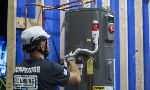 Photo of an adult man wearing a white safety helmet, gray gloves, and a black T‑shirt with the text “COMPETITOR” and “PLUMBING National Championship” on the back, installing a gray water heater connected to copper piping in an indoor construction setting.