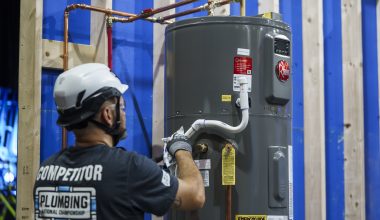 Photo of an adult man wearing a white safety helmet, gray gloves, and a black T‑shirt with the text “COMPETITOR” and “PLUMBING National Championship” on the back, installing a gray water heater connected to copper piping in an indoor construction setting.